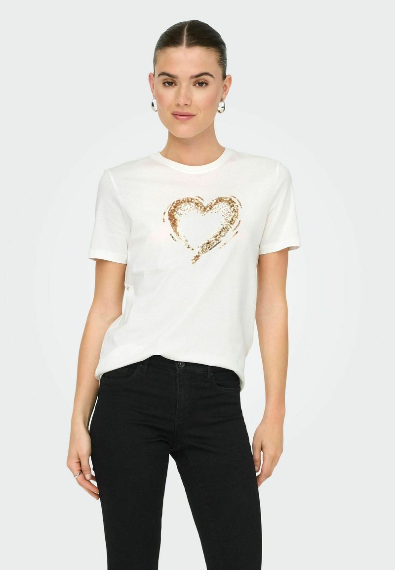 Camiseta blanca de algodón con un diseño de corazón hecho de lentejuelas doradas en la parte delantera. El modelo lleva jeans negros. La camiseta cuenta con cuello redondo y mangas cortas.