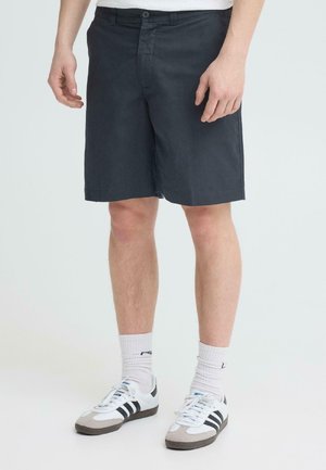 Mees, kes kannab musti põlvikuteni ulatuvaid lühikesi pükse, valgeid jalaümbriseid ja valgeid Adidas'i tossusid mustade triipudega, seisab ühevärvilise tausta ees.