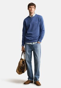Young man in blue sweatshirt, striped shirt, light blue jeans, brown shoes, holding a tan duffel bag with dark straps, standing against white background.