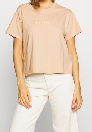 Kobieta w beżowym, krótkim t-shircie Levi's i białych spodniach z wysokim stanem, stoi z rękami swobodnie opuszczonymi wzdłuż ciała.