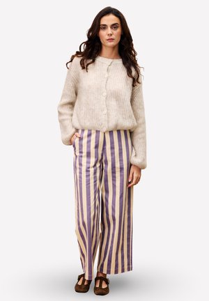 Vrouw in een beige knooptrui en paarse-beige verticaal gestreepte wijde broek, staande met één hand in de zak.