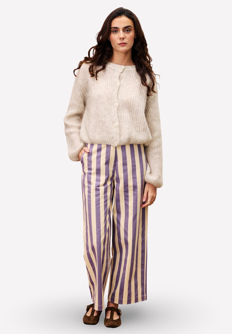 Vrouw in een beige knooptrui en paarse-beige verticaal gestreepte wijde broek, staande met één hand in de zak.