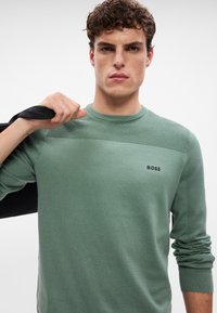 Zelený pletený sveter s rebrovanou textúrou na pleciach, okrúhlym výstrihom a malým čiernym logom "BOSS" na hrudi. Model drží čiernu tašku.