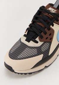Tenisky Nike Air Max majú vrchnú časť z vrstveného materiálu v čiernej farbe s mriežkovým vzorom, krémové a hnedé akcenty, textúrované prekrytia a čierne šnúrky.