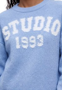 Pull bleu doux avec texte blanc duveteux "STUDIO 1993" sur la poitrine porté par une personne aux cheveux bouclés.