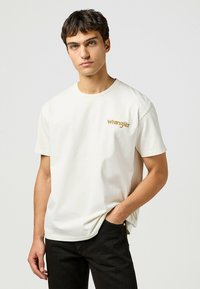Camiseta blanca de algodón con cuello redondo y mangas cortas, con un logo dorado de "Wrangler" en el lado izquierdo del pecho. Combinada con jeans de mezclilla negra.