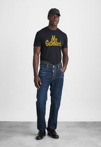 T-shirt noir avec texte jaune, jeans en denim bleu, et chaussures noires. Le modèle porte une casquette noire. Le décor est neutre, avec un fond gris clair.