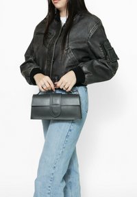 Borsa a tracolla in pelle grigia con texture liscia, forma rettangolare e chiusura a pattina, tenuta da una persona che indossa una giacca bomber nera e jeans chiari.