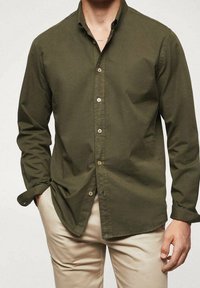 Camisa verde oliva de botones, con mangas largas, cuello y bolsillo frontal. Presenta botones blancos y una textura suave de algodón.
