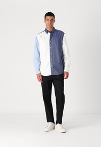 Chemise à boutons avec un design en blocs de couleurs en bleu foncé, bleu clair et blanc, en tissu de coton. Associée à un pantalon noir et des baskets blanches.