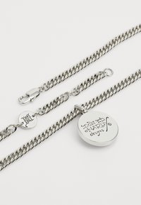 Sudraba metāla kaklarota ar vītne veida ķēdi un apaļu piekari, uz kura ir iegravēts "Don Ed Hardy designs." Iekļauta aizdare.