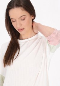 Top ușor, cu nervuri, de culoare crem, având mâneci lungi cu dungi roz și verde. Textură moale cu un decolteu relaxat.