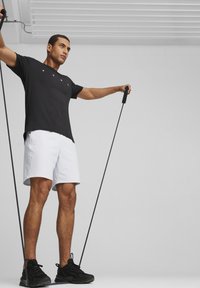 Camiseta atlética negra con logo, pantalones cortos blancos y zapatillas negras; sosteniendo bandas de resistencia con un fondo neutro.