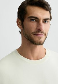 Lichtbeige sweatshirt met een geribbelde ronde hals, een soepele structuur en subtiele stikdetails, gedragen door een mannelijke model met kort haar.