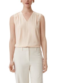 Mouwloze blouse van zacht perzikkleurig stof met een V-hals, met subtiele plooien bij de schouders. Gecombineerd met crèmekleurige broek.