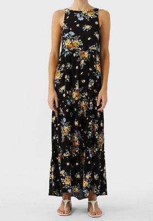 Czarna sukienka maxi bez rękawów z żółto-niebieskim kwiatowym wzorem, noszona z białymi płaskimi sandałami.