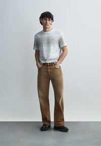 Γκρι πλεκτό μπλουζάκι με διακριτικό μοτίβο, σε συνδυασμό με καφέ τζιν παντελόνι ευθείας γραμμής. Το μοντέλο στέκεται μπροστά σε ουδέτερο φόντο. Μαύρα παπούτσια.