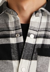 Zwart-wit geruit overhemd met metalen drukknoopjes, voorzien van een zachte textuur en een borstzak aan de linkerkant.