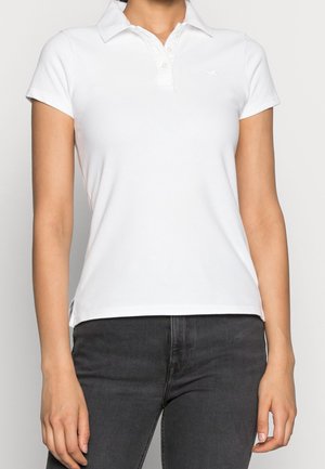 Femme portant un polo blanc cintré à manches courtes avec des boutons et un jean gris foncé, debout devant un fond uni.