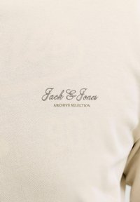 Tissu beige en gros plan avec texte brodé gris "Jack & Jones" et texte doré plus petit "ARCHIVE SELECTION" sur le vêtement.