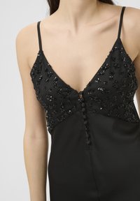 Crna večernja haljina s v-izrezom i ukrasima od perlica na korpici; ima tanke naramenice i dekorativne dugmadi duž prednje strane.