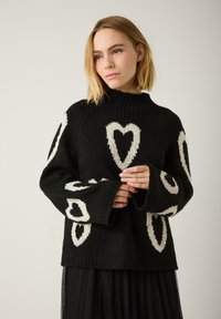 Schwarzer gestrickter Pullover mit weißen Herzmustern, hohem Kragen und weiten Ärmeln. Ausgestattet mit einem strukturierten, groben Strickfinish.