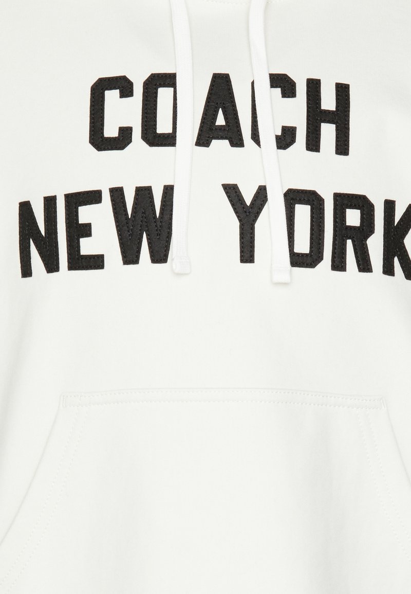 Witte hoodie met zwart geborduurde tekst "COACH NEW YORK" op de borst, met trekkoorden en een front pocket. Zacht materiaal.