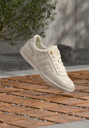 Zapatilla blanca de Tommy Hilfiger flotando sobre un suelo de madera con baldosas, con vegetación y una pared de hormigón al fondo.