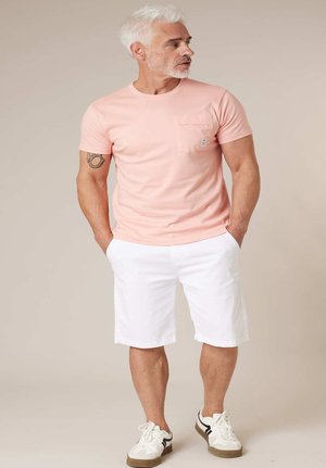 Homme aux cheveux blancs et à la barbe portant une chemise rose à manches courtes, un short blanc jusqu'aux genoux et des baskets blanches, debout les mains dans les poches.