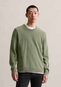 Zielony sweter z dzianiny z okrągłym dekoltem i ribb Nożami. Założony na białą koszulę, z fakturowanym wzorem. Komponuje się z czarnymi dżinsami.