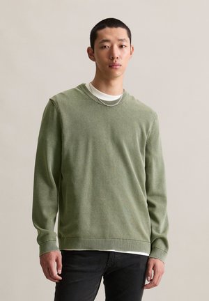 Green knit sweater featuring a round neckline and ribbed cuffs. Layered over a white shirt, with a textured pattern. Complements black jeans.