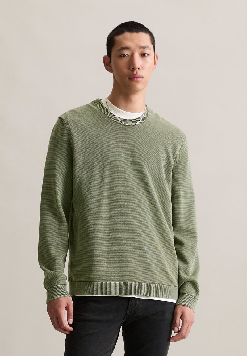 Zielony sweter z dzianiny z okrągłym dekoltem i ribb Nożami. Założony na białą koszulę, z fakturowanym wzorem. Komponuje się z czarnymi dżinsami.