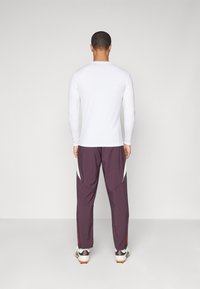 Witte sportshirt met lange mouwen in combinatie met paarse broek met witte accenten en rode rand, gezien vanuit de achterkant.