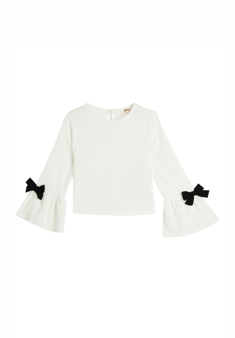 Top bianco a maniche lunghe con costine e maniche a campana, caratterizzato da dettagli in nero a forma di fiocco ai polsini. Chiusura sul retro del collo.