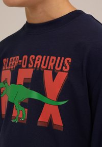 Mornarsko modra bombažna majica z dolgimi rokavi, ki prikazuje besedilo "SLEEP-O SAURUS REX" v rdeči barvi in z zeleni dinozaverjem, z okroglim izrezom.