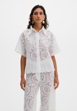 Frau mit lockigem Haar trägt ein weißes, florales Spitzen-Shortsleeve-Hemd mit Knöpfen und passende weit geschnittene Hosen, dazu große goldene Muschel-Ohrringe als Accessoire.