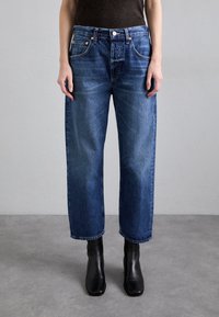 Blå denimjeans med rak passform, femficksdesign och synlig sömnad. Bärs med svarta ankelstövlar, mot en neutral bakgrund.