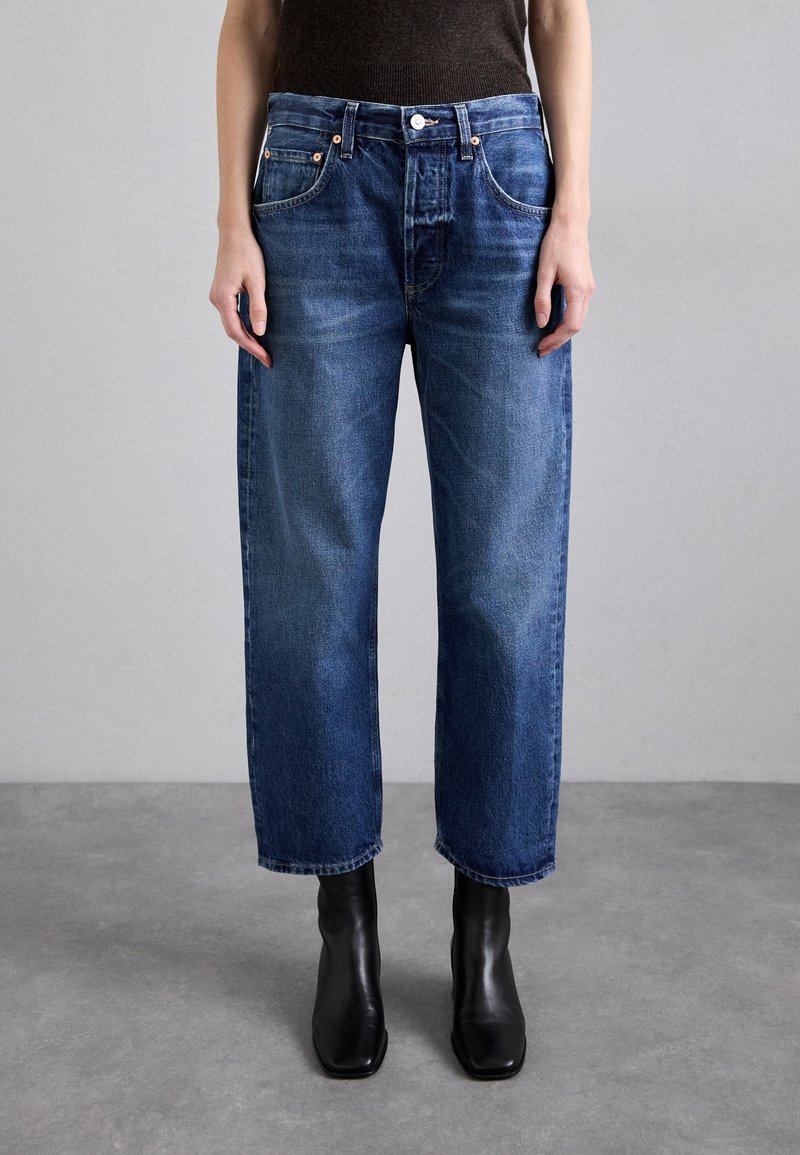 Blå denimjeans med rak passform, femficksdesign och synlig sömnad. Bärs med svarta ankelstövlar, mot en neutral bakgrund.