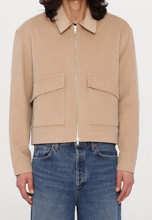 Beżowa kurtka sztruksowa z przednim zamkiem błyskawicznym i dużymi kieszeniami z patkami, noszona z niebieskimi dżinsami.