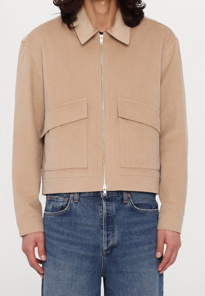Veste beige en velours côtelé avec zip frontal et grandes poches à rabat, portée avec un jean bleu.