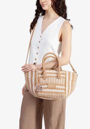 Donna con capelli ricci che indossa una canottiera bianca con bottoni e pantaloni marroni, che tiene in mano una borsa intrecciata color beige e bianco con doppio manico.