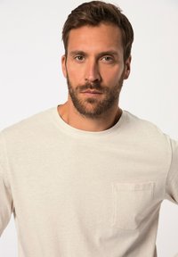 Helle beige Langarmshirt mit Rundhalsausschnitt und Brusttasche. Der Stoff wirkt weich mit einer dezenten Textur.