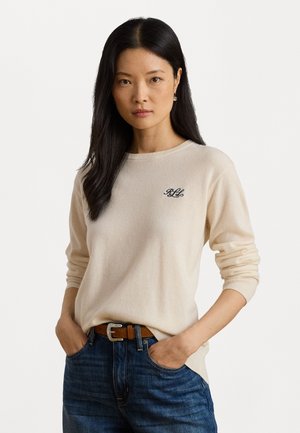 Beige knit sweater with a rounded neckline and ribbed cuffs, featuring a small embroidered logo on the chest; paired with dark jeans and a brown belt.