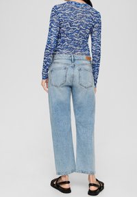 Kvinna i blå camouflage långärmad tröja, ljusblå högmidjade raka jeans och svarta sandaler, stående med ryggen vänd mot kameran.