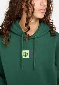 Sudadera verde hecha de material suave con un gráfico frontal que presenta dos emblemas geométricos en verde claro y púrpura oscuro. Cuerda ajustable.