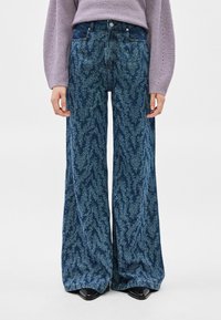 Γυναίκα που φοράει μπλε τζιν με ευρύ πόδι και σχέδιο, λιλά πλεκτό πουλόβερ και μαύρα μυτερά παπούτσια, εμφανιζόμενη από τη μέση έως τα πόδια.