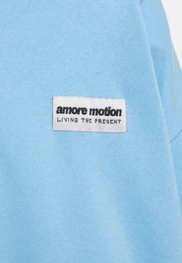 Svetlomodrá bavlnená tkanina s bielou etikettou a čiernym textom, ktorý znie "amore motion" a "ŽIť prítomnosťou". Jednoduchý dizajn bez vzorov.