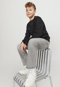 Felpa nera con testo blu, jeans grigi e sneaker bianche. Seduto su una sedia in metallo argentato con un design semplice e a righe.