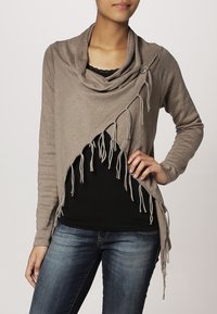 Cardigan avvolgente beige con colletto drappeggiato, maniche lunghe e dettagli frangiati sui lati. Indossato sopra un top nero e abbinato a jeans blu.
