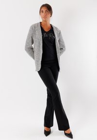 Blazer grigio texturizzato con cappuccio, indossato sopra una maglietta nera. I pantaloni neri a campana e i décolleté neri a punta completano l'outfit.